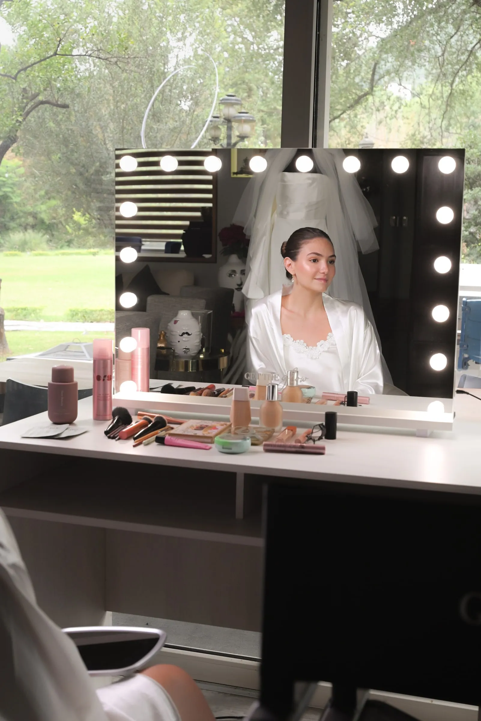 Novia reflejada en el espejo Hollywood mientras la maquillan, con los productos sobre la mesa