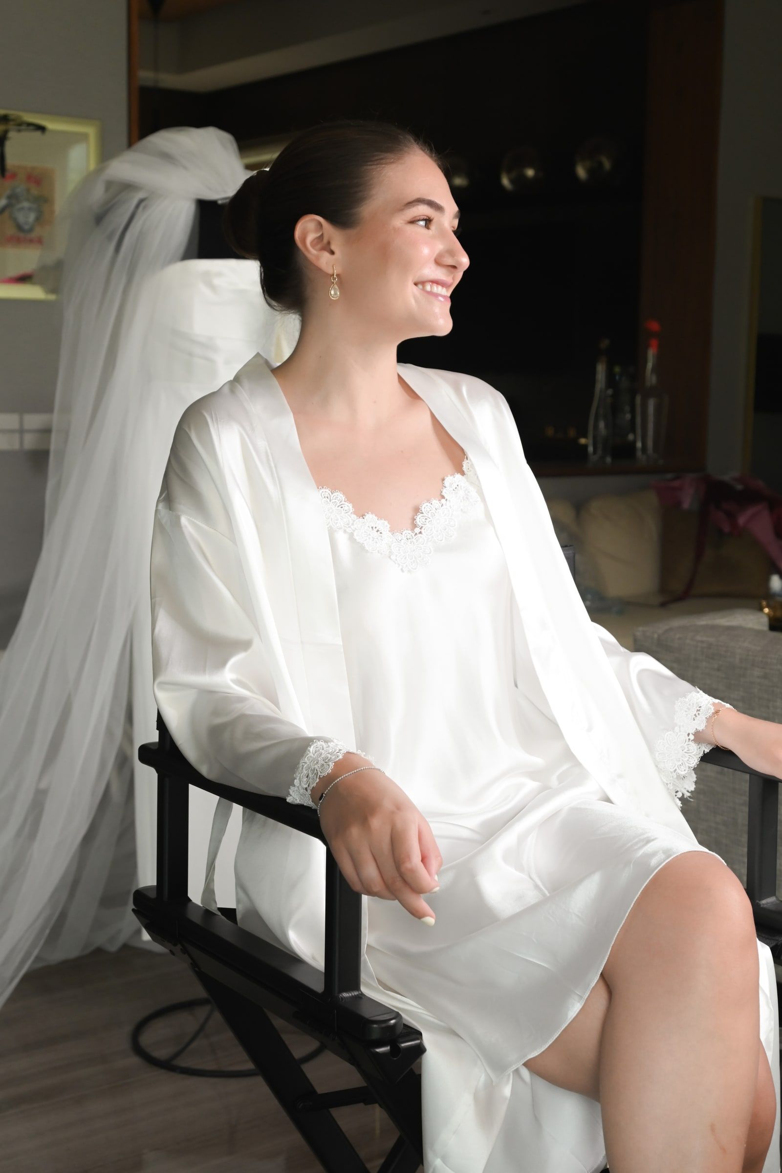 Novia en bata blanca sentada en la silla de estilista, riendo, con el velo colgado detrás